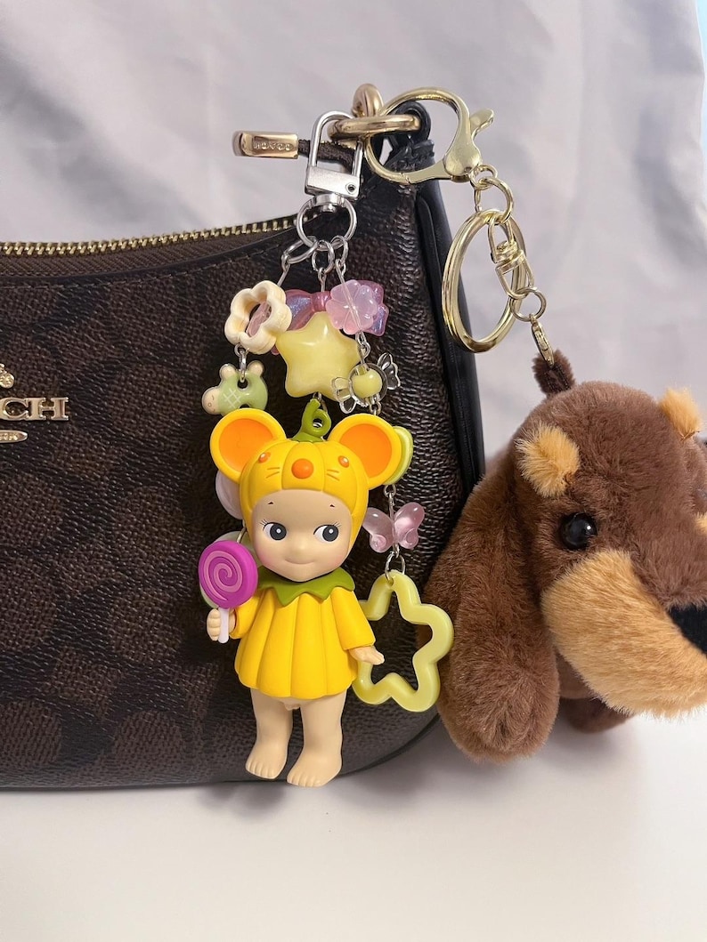Puede incluir: Un llavero decorativo con una figura amarilla con disfraz de rat&oacute;n sosteniendo una piruleta, un oso de peluche marr&oacute;n y varios abalorios. El llavero est&aacute; sujeto a un bolso marr&oacute;n con herrajes dorados.