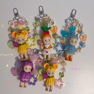 Puede incluir: Cinco llaveros con peque&ntilde;as figuras disfrazadas, incluyendo una calabaza, una zanahoria y un rat&oacute;n. Cada llavero tiene una cadena de cuentas decorativa con abalorios, estrellas y corazones. Las figuras son de color amarillo, naranja, morado y azul.