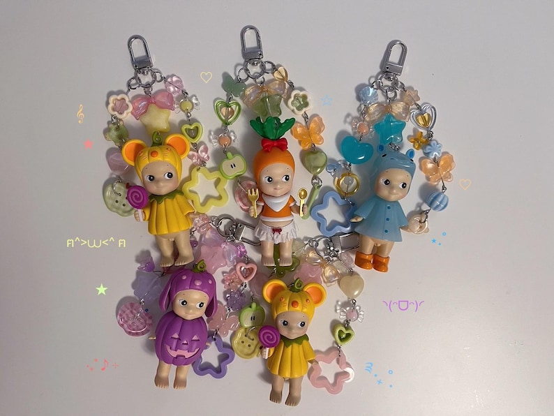 Puede incluir: Cinco llaveros con peque&ntilde;as figuras disfrazadas, incluyendo una calabaza, una zanahoria y un rat&oacute;n. Cada llavero tiene una cadena de cuentas decorativa con abalorios, estrellas y corazones. Las figuras son de color amarillo, naranja, morado y azul.