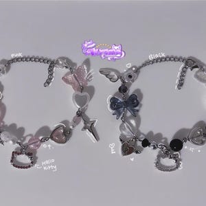Op de afbeelding: Twee armbanden met kralen, één roze en één zwart, met bedels waaronder vlinders, harten, strikken en Hello Kitty-hangers. De armbanden zijn verbonden door een zilveren ketting.