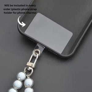 Puede incluir: Primer plano de una funda de tel&eacute;fono negra con un soporte de correa de pl&aacute;stico transparente. Un cierre plateado conecta la correa a un cord&oacute;n de cuentas con cuentas blancas transl&uacute;cidas. Texto: "Will be included in every order..."