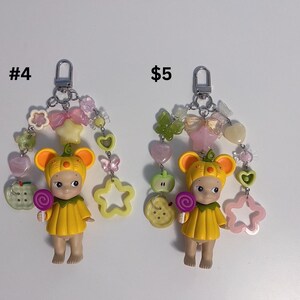 Puede incluir: Dos llaveros con una peque&ntilde;a mu&ntilde;eca con un atuendo y sombrero amarillos, sosteniendo una piruleta. Cada llavero tiene un cierre plateado y est&aacute; adornado con cuentas de colores, dijes y un dije en forma de estrella. Los textos "#4" y "$5" son visibles.