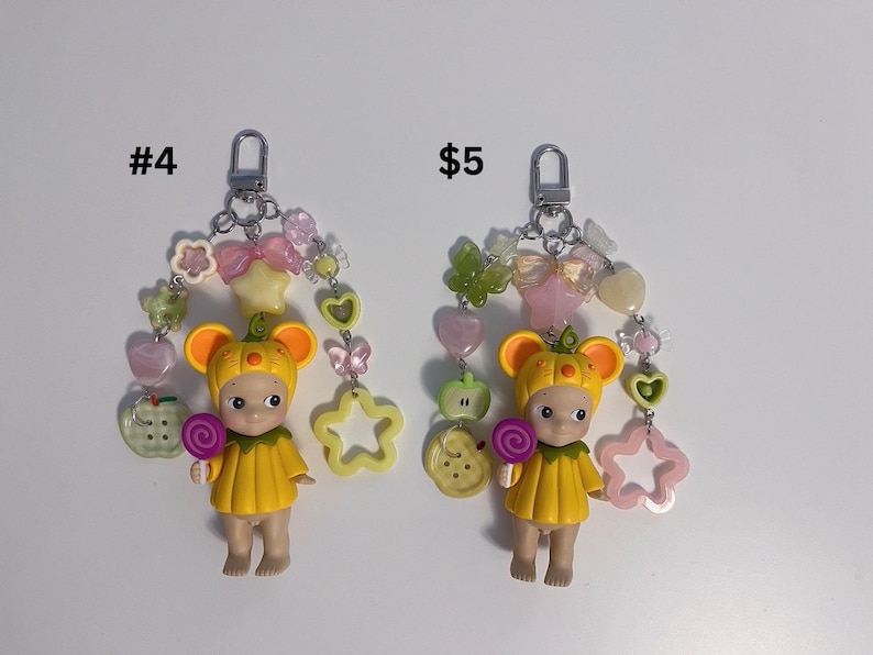 Puede incluir: Dos llaveros con una peque&ntilde;a mu&ntilde;eca con un atuendo y sombrero amarillos, sosteniendo una piruleta. Cada llavero tiene un cierre plateado y est&aacute; adornado con cuentas de colores, dijes y un dije en forma de estrella. Los textos "#4" y "$5" son visibles.