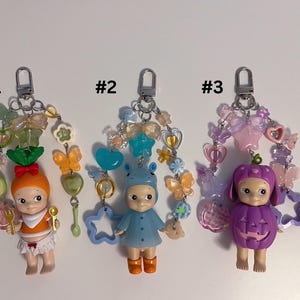 Puede incluir: Tres llaveros con peque&ntilde;as figuras de mu&ntilde;ecas. La primera mu&ntilde;eca viste un atuendo naranja y tiene una zanahoria en la cabeza. La segunda mu&ntilde;eca viste un atuendo azul con un gorro de oso. La tercera mu&ntilde;eca est&aacute; vestida como una calabaza morada. Cada llavero tiene cuentas decorativas.