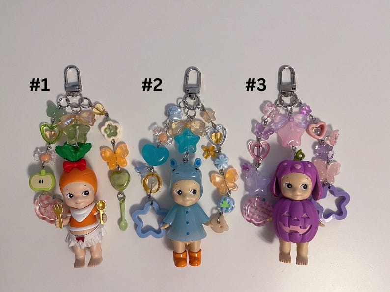 Puede incluir: Tres llaveros con peque&ntilde;as figuras de mu&ntilde;ecas. La primera mu&ntilde;eca viste un atuendo naranja y tiene una zanahoria en la cabeza. La segunda mu&ntilde;eca viste un atuendo azul con un gorro de oso. La tercera mu&ntilde;eca est&aacute; vestida como una calabaza morada. Cada llavero tiene cuentas decorativas.
