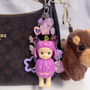 Puede incluir: Un llavero morado con tem&aacute;tica de calabaza con una cara sonriente, sujeto a un bolso marr&oacute;n. El llavero est&aacute; adornado con cuentas rosas y moradas, estrellas y un coraz&oacute;n. Tambi&eacute;n hay un peque&ntilde;o perro de peluche marr&oacute;n adjunto.