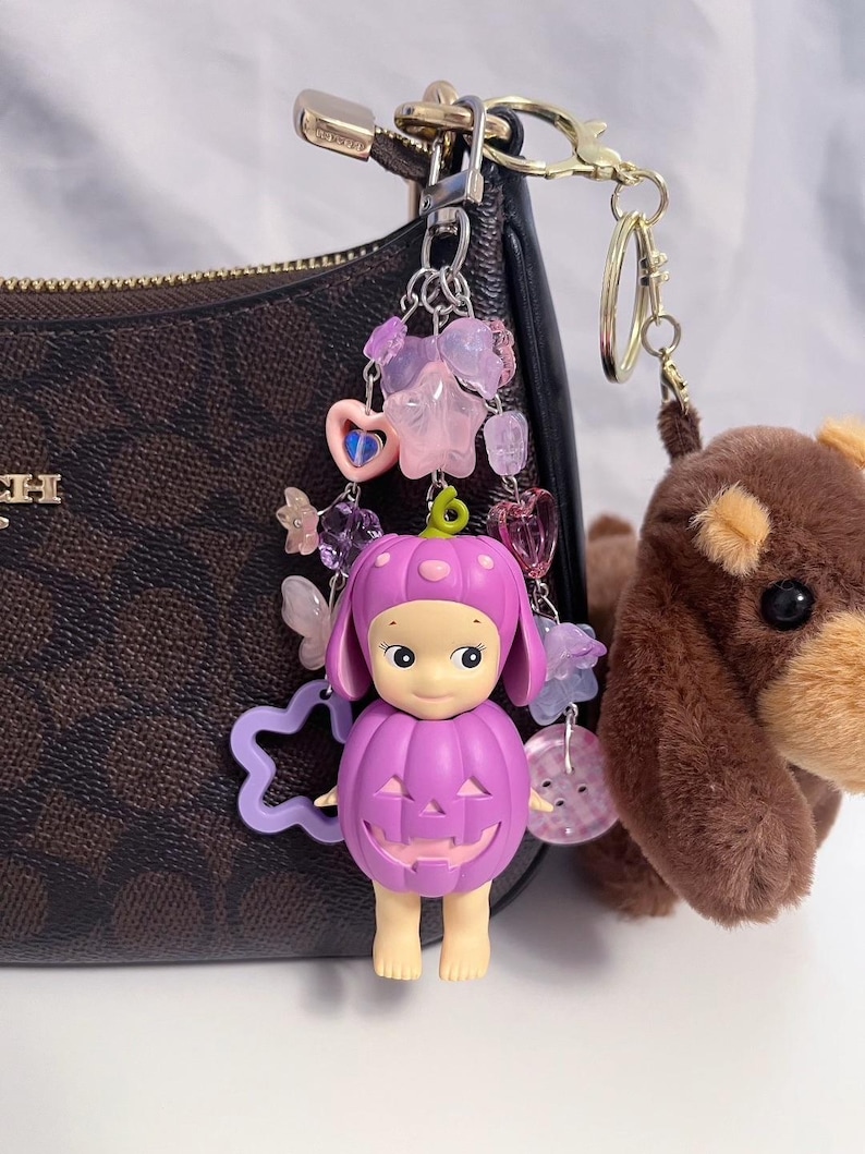 Puede incluir: Un llavero morado con tem&aacute;tica de calabaza con una cara sonriente, sujeto a un bolso marr&oacute;n. El llavero est&aacute; adornado con cuentas rosas y moradas, estrellas y un coraz&oacute;n. Tambi&eacute;n hay un peque&ntilde;o perro de peluche marr&oacute;n adjunto.