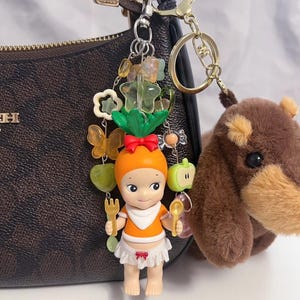 Puede incluir: Un llavero decorativo con una peque&ntilde;a mu&ntilde;eca con sombrero y top naranja, sosteniendo un tenedor y una cuchara. El llavero incluye dijes de mariposas, flores y frutas, junto con un oso de peluche marr&oacute;n. La mu&ntilde;eca lleva un babero y una falda blancos.
