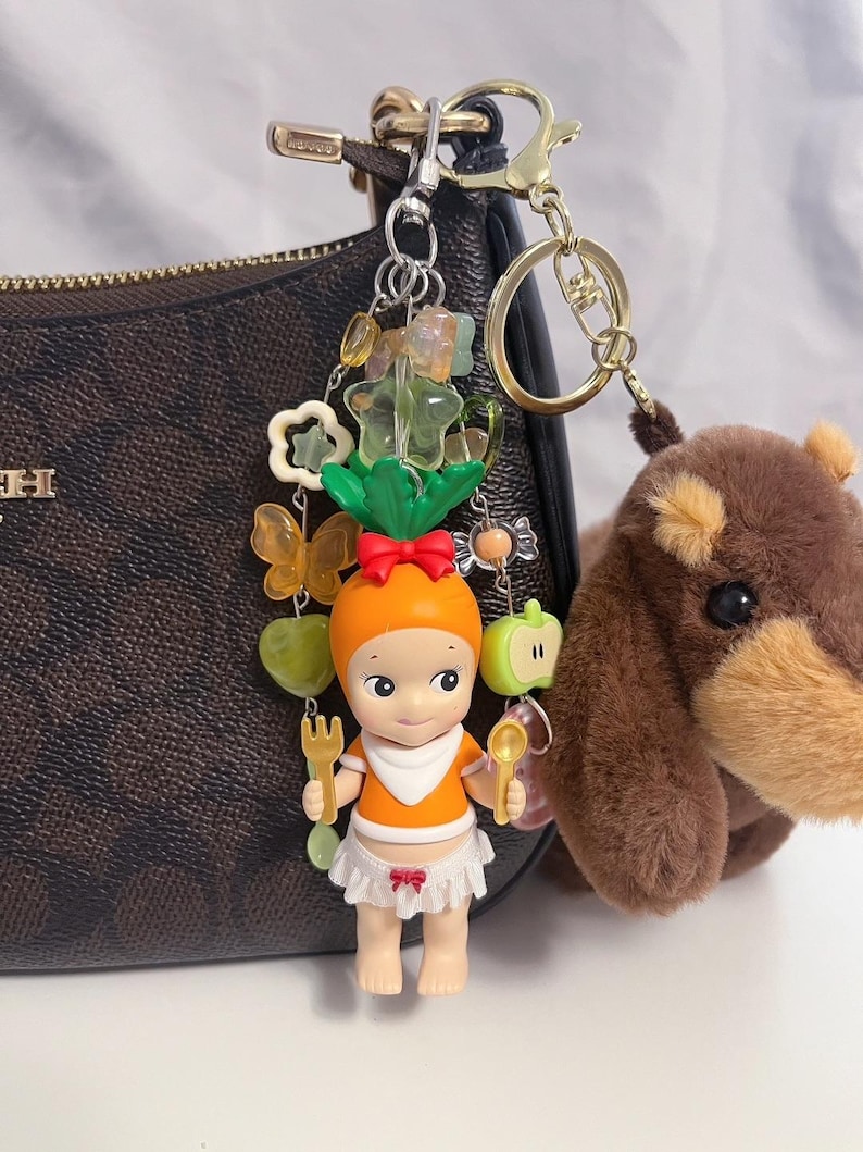 Puede incluir: Un llavero decorativo con una peque&ntilde;a mu&ntilde;eca con sombrero y top naranja, sosteniendo un tenedor y una cuchara. El llavero incluye dijes de mariposas, flores y frutas, junto con un oso de peluche marr&oacute;n. La mu&ntilde;eca lleva un babero y una falda blancos.