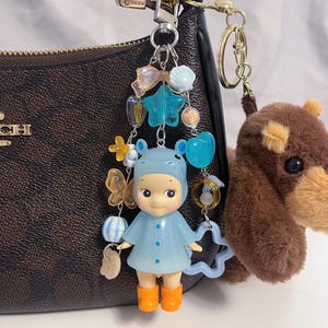 Puede incluir: Un llavero de mu&ntilde;eca azul con botas naranjas y un abrigo azul a juego, adornado con abalorios de estrella, cuentas y lazos. Un llavero de perro de peluche marr&oacute;n cuelga cerca. Los llaveros est&aacute;n sujetos a un bolso marr&oacute;n con herrajes dorados.