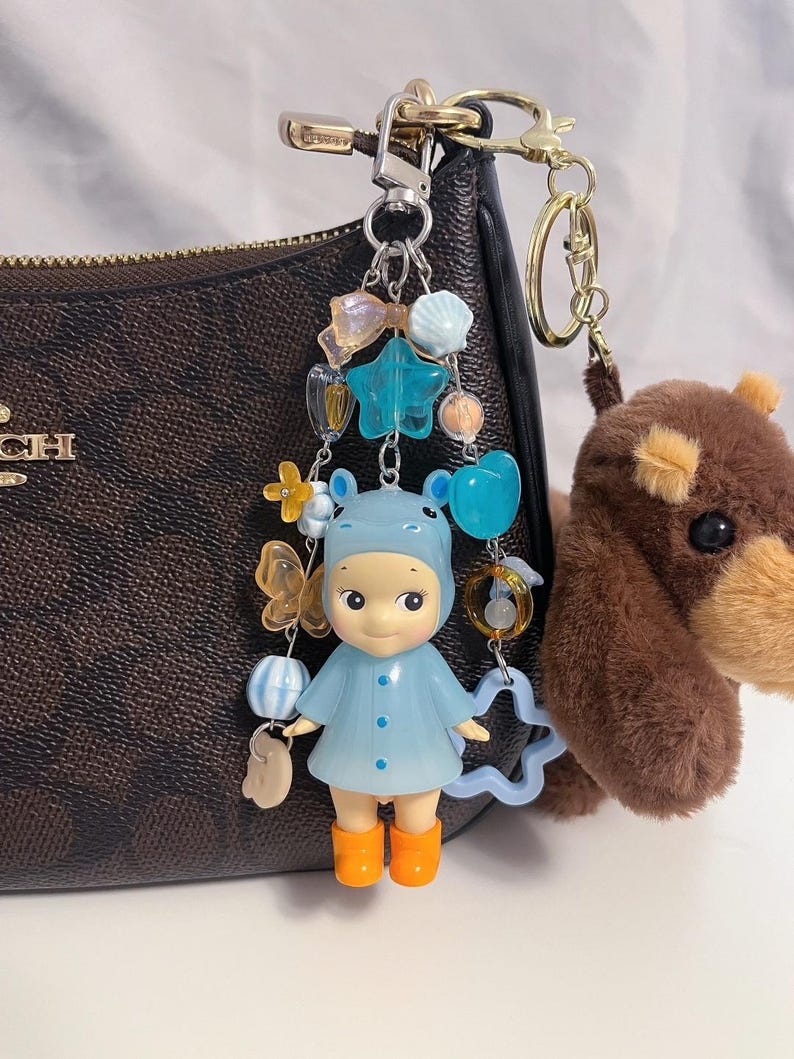 Puede incluir: Un llavero de mu&ntilde;eca azul con botas naranjas y un abrigo azul a juego, adornado con abalorios de estrella, cuentas y lazos. Un llavero de perro de peluche marr&oacute;n cuelga cerca. Los llaveros est&aacute;n sujetos a un bolso marr&oacute;n con herrajes dorados.