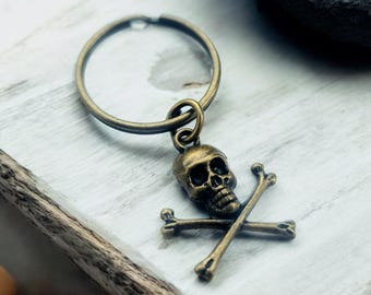 May include: A bronze skull and crossbones keychain. The skull is detailed and the crossbones are simple. The keychain is attached to a bronze ring.