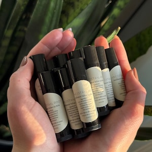 May include: A close-up of a hand holding multiple black tubes of lip balm. The tubes are labeled "Ancestral Root" and "Tallow Lip Balm".