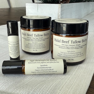 Könnte beinhalten: Drei Glasbehälter mit schwarzen Deckeln, die Hautpflegeprodukte enthalten. Die Behälter sind mit "Halal Beef Tallow Balm" und "Age Rewind Oil Roller" beschriftet. Die Behälter stehen auf einer weißen Oberfläche.