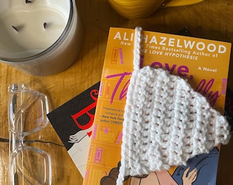 May include: A white crocheted bookmark rests on a book titled "The Love Hypothesis" by Ali Hazelwood. A yellow mug of coffee, a candle, and eyeglasses are also on the wooden surface.
