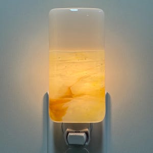 May include: A rectangular glass night light plugged into a wall outlet. The upper portion is opaque white, while the lower section features a swirling, marbled pattern of translucent amber, gold, and cream tones that glow when illuminated.