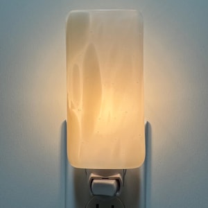 May include: A rectangular plug-in night light featuring a translucent, frosted glass cover with a soft, warm yellow glow. The glass surface displays subtle, cloudy white swirls and small air bubbles throughout, mounted on a white plastic base.