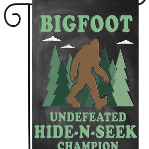 May include: A black and green garden flag with a silhouette of Bigfoot walking through a forest. The text reads "Bigfoot Undefeated Hide-N-Seek Champion."