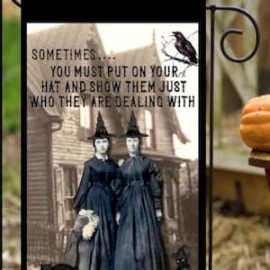 May include: A black and white garden flag with a quote about witches: "Sometimes... you must put on your hat and show them just who they are dealing with." Two witches in black dresses and hats stand in front of a house with a black crow perched on a branch.