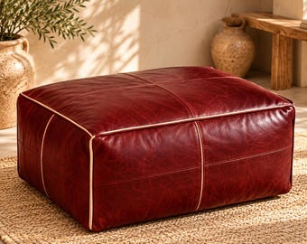 Può includere: Un pouf rettangolare rivestito in pelle rosso scuro con una texture a grana visibile. Il pezzo presenta un profilo di colore chiaro prominente lungo i bordi e le cuciture, creando un motivo a griglia sulla parte superiore e sui lati.