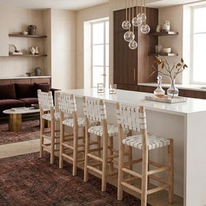 May include: Four bar stools with light-toned wooden frames and white woven leather seats and backrests. Each stool features a sturdy four-legged base with horizontal support rungs, positioned in a row along a white kitchen island countertop.