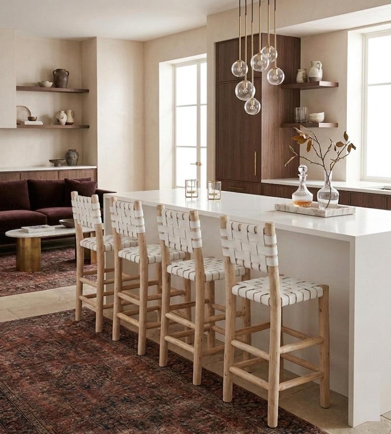 May include: Four bar stools with light-toned wooden frames and white woven leather seats and backrests. Each stool features a sturdy four-legged base with horizontal support rungs, positioned in a row along a white kitchen island countertop.
