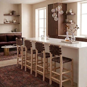 May include: Four bar stools with light-toned wooden frames and dark brown woven leather seats and backrests are positioned along a white kitchen island. The stools feature a tall, sturdy design with horizontal footrests and a simple, clean structure.