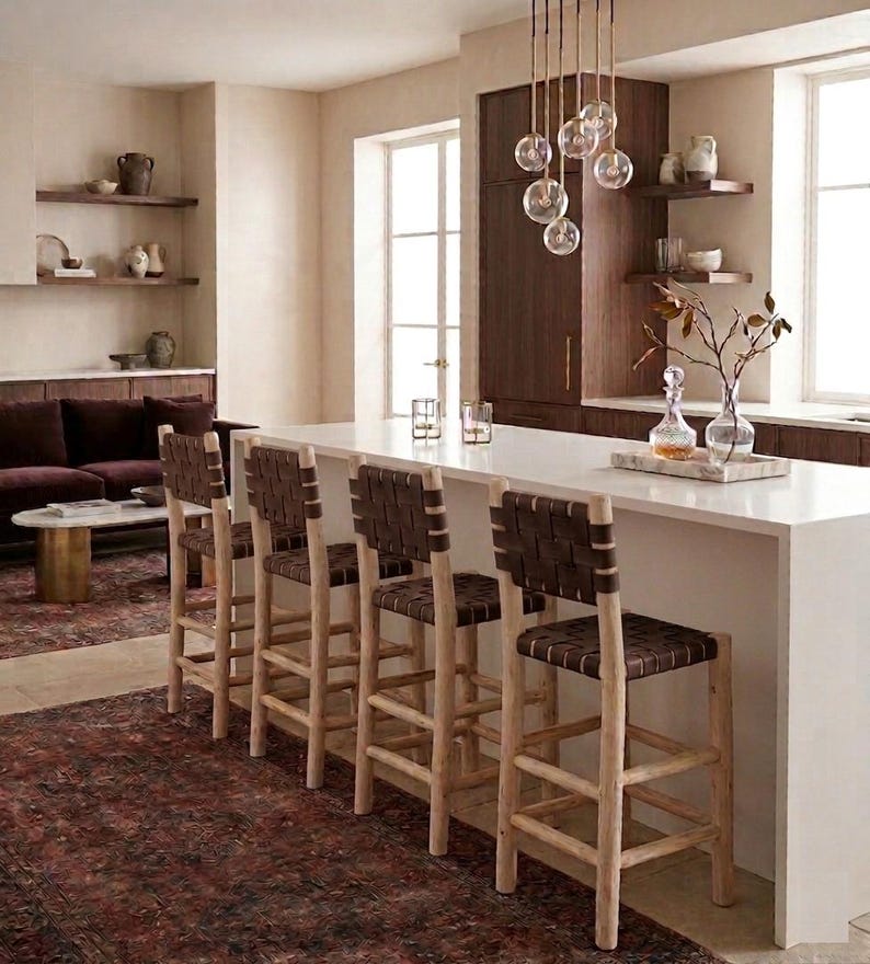 May include: Four bar stools with light-toned wooden frames and dark brown woven leather seats and backrests are positioned along a white kitchen island. The stools feature a tall, sturdy design with horizontal footrests and a simple, clean structure.