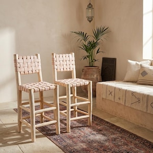 May include: Two bar stools featuring light-colored wooden frames with four legs and horizontal footrests. The seats and backrests are constructed from woven light tan leather straps in a grid pattern, providing a structured and supportive seating surface.