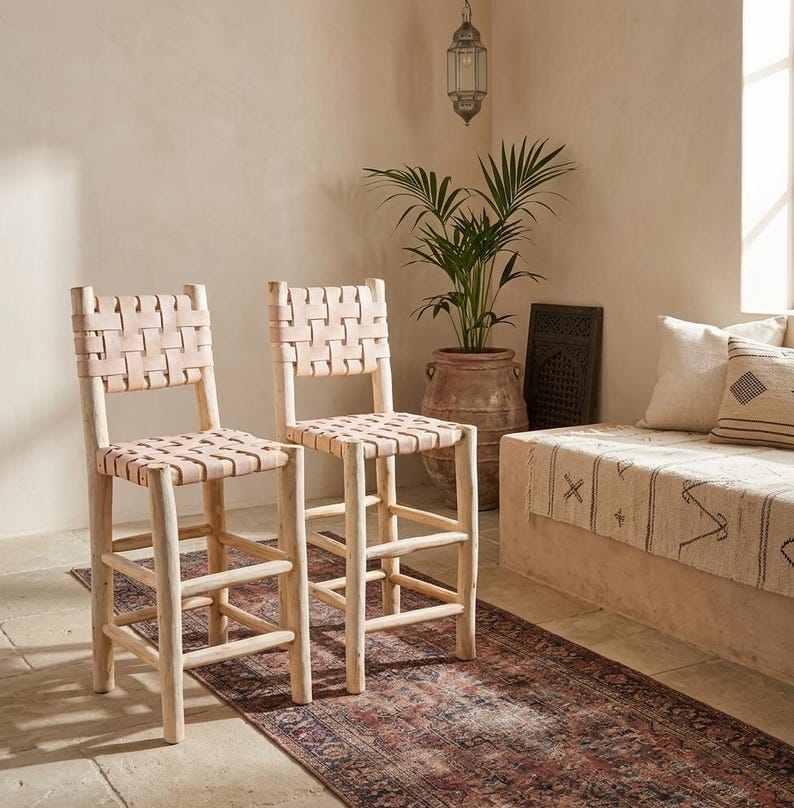 May include: Two bar stools featuring light-colored wooden frames with four legs and horizontal footrests. The seats and backrests are constructed from woven light tan leather straps in a grid pattern, providing a structured and supportive seating surface.