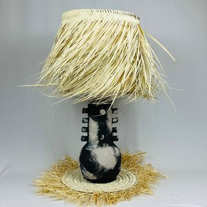 Peut inclure: Une lampe unique avec une esthétique naturelle et bohème. La lampe présente une base cylindrique avec un motif peint en noir et blanc, surmontée d'un abat-jour en paille tressée. La lampe repose sur un tapis rond en paille.
