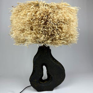 K&ouml;nnte beinhalten: Eine einzigartige Tischlampe mit einem strukturierten, beigefarbenen Lampenschirm aus Naturfasern. Der Sockel hat eine dunkle, organische Form mit einem Ausschnitt, und die Lampe hat ein schwarzes Kabel und einen Schalter. Diese dekorative Lampe.