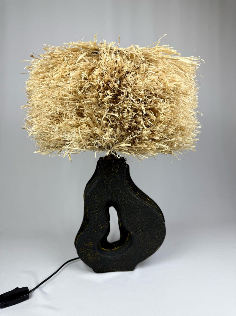 K&ouml;nnte beinhalten: Eine einzigartige Tischlampe mit einem strukturierten, beigefarbenen Lampenschirm aus Naturfasern. Der Sockel hat eine dunkle, organische Form mit einem Ausschnitt, und die Lampe hat ein schwarzes Kabel und einen Schalter. Diese dekorative Lampe.