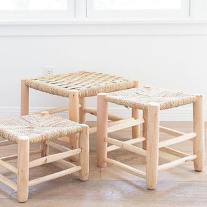 May include: Three natural woven wicker ottomans with a light wood frame. The ottomans are arranged in a staggered formation on a light wood floor.