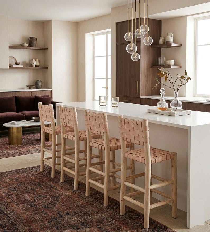 May include: Four bar stools with light-colored wooden frames and seats and backrests featuring a woven tan leather strap design. The stools are positioned along a white kitchen island countertop in a room with dark wood cabinetry and decor.