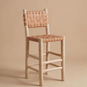 May include: A bar stool featuring a light-colored wooden frame with four cylindrical legs and horizontal support rungs. The seat and backrest are constructed from tan leather strips woven in a grid pattern, providing a structured surface.