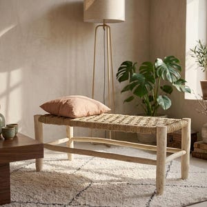 Può includere: Panca in legno chiaro con seduta intrecciata e cuscino rosa. La panca è in una stanza con un tappeto, piante e una lampada da terra. Un tavolino in legno scuro contiene una teiera e tazze.