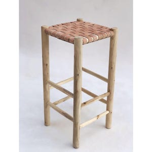 May include: A tall, wooden stool with a woven leather seat. The stool has four sturdy legs and horizontal supports for stability. The seat is a light brown color, and the wood has a natural, unfinished appearance. The stool is designed for seating.