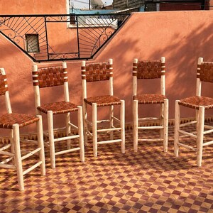 May include: Five tall bar stools featuring light-colored natural wood frames with four legs and horizontal support rungs. Each stool has a square seat and a rectangular backrest constructed from woven brown leather strips in a basket-weave pattern.