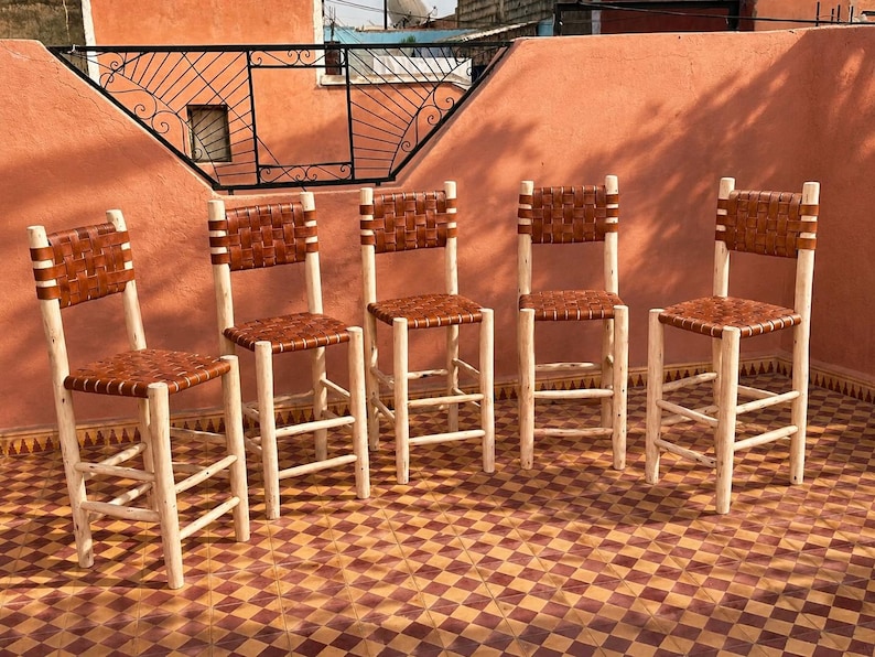 May include: Five tall bar stools featuring light-colored natural wood frames with four legs and horizontal support rungs. Each stool has a square seat and a rectangular backrest constructed from woven brown leather strips in a basket-weave pattern.
