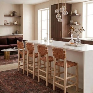 May include: Four bar stools with light-colored wooden frames and woven tan leather seats and backrests. The stools are positioned along a white kitchen island. Each stool features a sturdy four-legged base with horizontal support rungs.