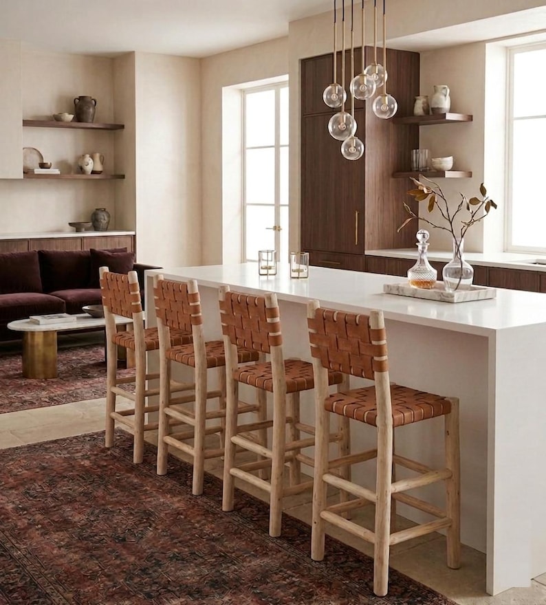 May include: Four bar stools with light-colored wooden frames and woven tan leather seats and backrests. The stools are positioned along a white kitchen island. Each stool features a sturdy four-legged base with horizontal support rungs.