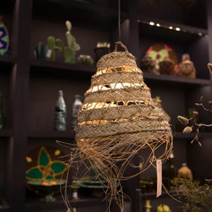 May include: A woven, light brown, pendant lampshade with a fringe of natural fibers. The lampshade is hanging from a ceiling in a room with shelves and other decorative items.