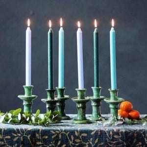 Peut inclure: Six bougies coniques allumées de différentes nuances de bleu et de vert, placées dans des bougeoirs en céramique verte. Les bougies sont disposées sur une nappe à motifs, avec de la verdure et des oranges.