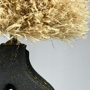 K&ouml;nnte beinhalten: Dekorative Lampe mit einem schwarzen Sockel mit rissiger, strukturierter Oberfl&auml;che und goldenen Akzenten. Der Lampenschirm besteht aus einem dichten, naturfarbenen, stroh&auml;hnlichen Material, das eine einzigartige und strukturierte &Auml;sthetik erzeugt. Die Lampe ist ein dekorativer Einrichtungsgegenstand.