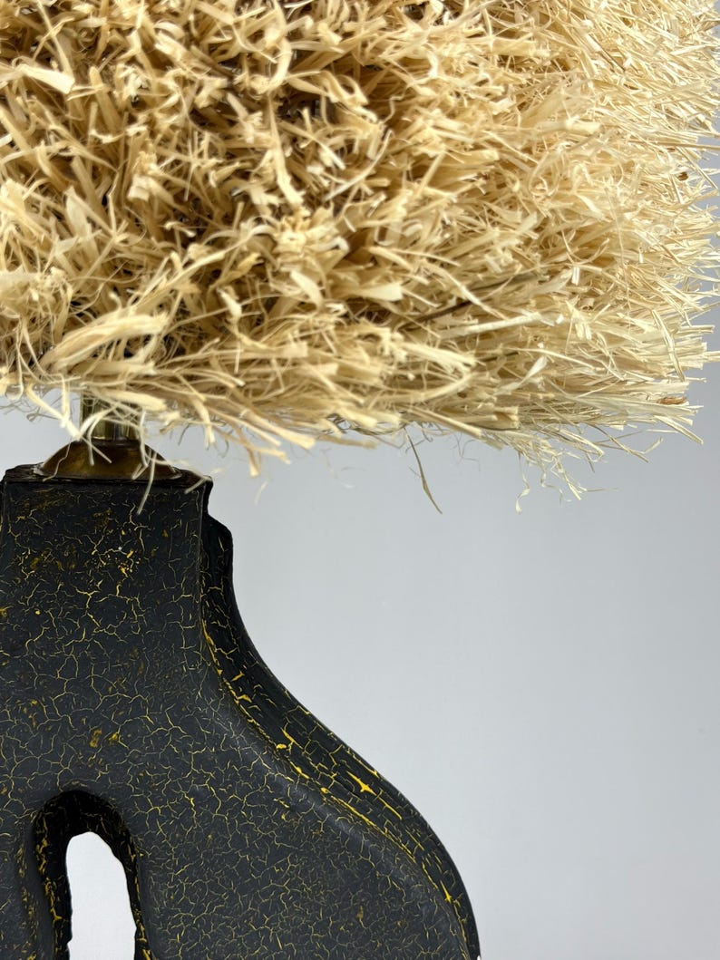 K&ouml;nnte beinhalten: Dekorative Lampe mit einem schwarzen Sockel mit rissiger, strukturierter Oberfl&auml;che und goldenen Akzenten. Der Lampenschirm besteht aus einem dichten, naturfarbenen, stroh&auml;hnlichen Material, das eine einzigartige und strukturierte &Auml;sthetik erzeugt. Die Lampe ist ein dekorativer Einrichtungsgegenstand.