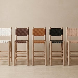 May include: Five bar stools with light-colored wooden frames and woven seats and backrests. The stools feature different colored woven straps in white, dark brown, tan, black, and light beige, each with a square back and four legs with rungs.
