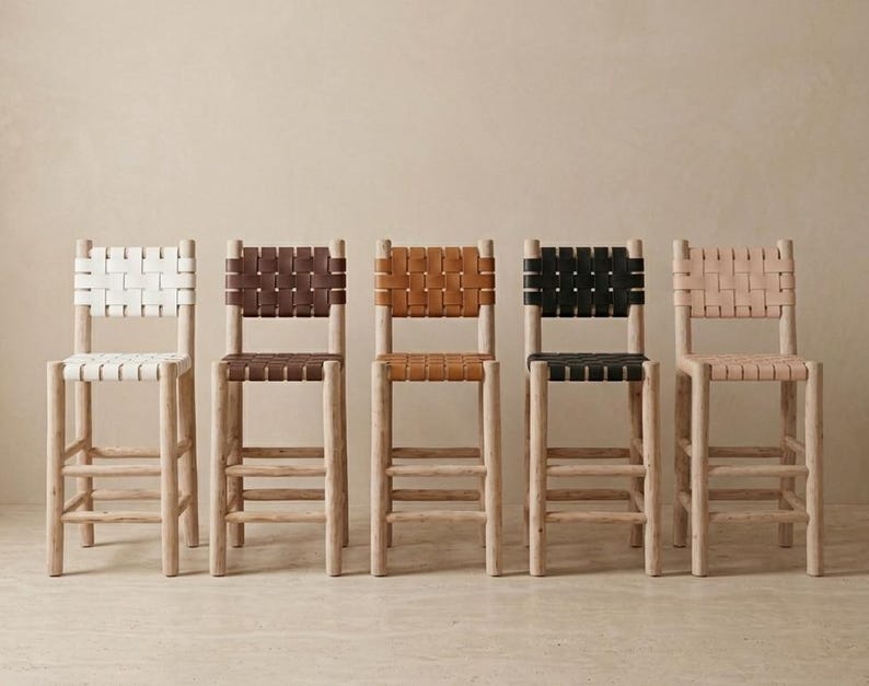 May include: Five bar stools with light-colored wooden frames and woven seats and backrests. The stools feature different colored woven straps in white, dark brown, tan, black, and light beige, each with a square back and four legs with rungs.