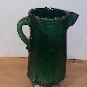 May include: A handcrafted, dark green ceramic jug with a handle and spout. The jug has a glossy finish and decorative detailing around the rim and base. The jug is approximately 18 cm tall.