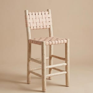 May include: A bar stool featuring a light-colored wooden frame with four cylindrical legs and horizontal support rungs. The seat and backrest are constructed from wide, light beige leather strips woven together in a simple grid pattern.